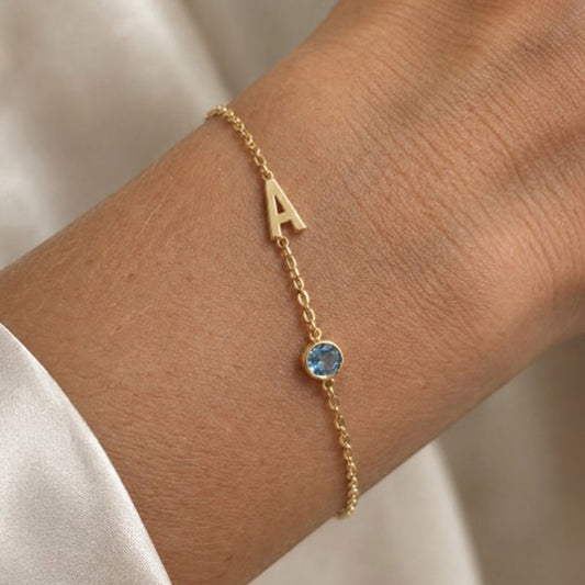 A delicate gold chain bracelet worn on a wrist, featuring a gold letter 'A' and a small round aquamarine birthstone, shown against a soft neutral background.